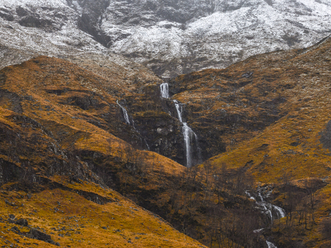 Aerial view of a waterfall cascading down rugged mountain slopes with snow dusted peaks and autumnal vegetation in Glencoe Scotland United Kingdom. Aerial view of a waterfall cascading down rugged mountain slopes with snow dusted peaks and autumnal vegetation in Glencoe Scotland United Kingdom.