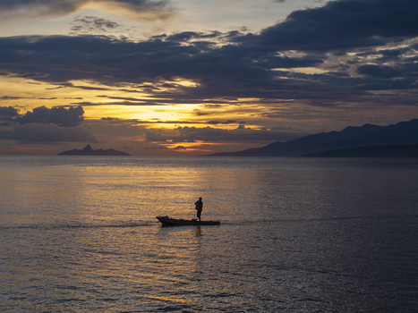 Aerial view of a lone fisherman in a small wooden boat on the calm sea at sunset with distant mountains under a dramatic golden sky in Flores Indonesia.