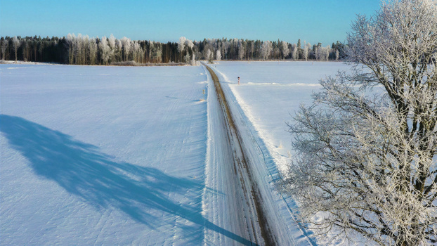 Aerial view of a straight road cutting through a vast snow covered landscape with frost covered trees under a clear blue sky in Latvia. Aerial view of a straight road cutting through a vast snow covered landscape with frost covered trees under a clear blue sky in Latvia.