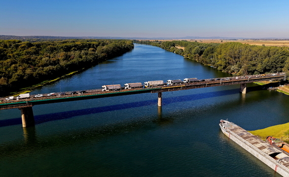 Aerial view of the bridge over the Danube River with trucks crossing and a barge moored below surrounded by lush riverbanks under a clear blue sky in Smederevo Podunavlje District Serbia.