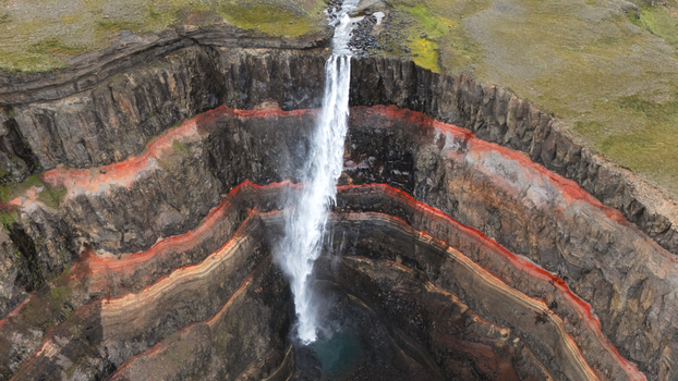 Aerial view of Hengifoss waterfall cascading down the cliff face with striking red clay layers contrasting against the dark basalt rock Iceland.