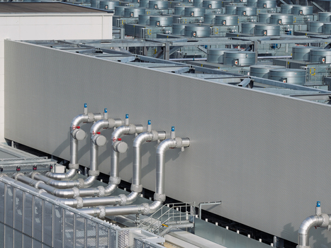 Aerial view of a data center roof featuring rows of industrial cooling fans and metallic piping systems in the United Kingdom. Aerial view of a data center roof featuring rows of industrial cooling fans and metallic piping systems in the United Kingdom.