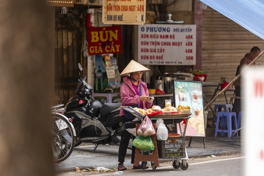 Hanoi Vietnam 11 November 2025 View of a street food vendor in a conical hat preparing snacks at a mobile cart on a city street with a motorbike and restaurant signage.