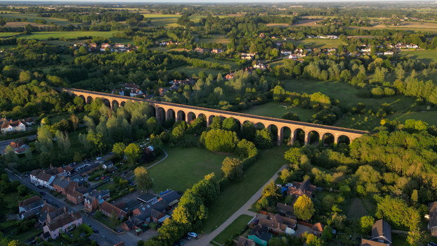 Aerial view of the historic Chappel Viaduct spanning the lush green landscape with surrounding trees rural houses and golden hour sunlight in Chappel England United Kingdom.