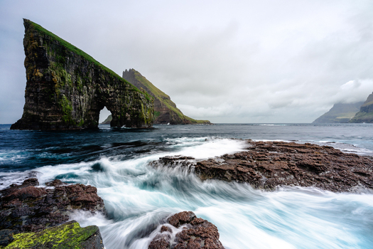 View of Drangarnir sea stacks with crashing waves against the rocky coastline under an overcast sky in the Faroe Islands.