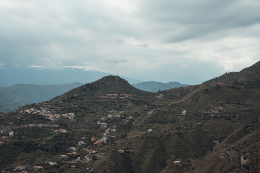 View of rugged verdant mountains dotted with quaint settlements under a vast moody sky creating a contrasting landscape Taormina Sicily Italy.