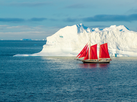 Aerial view of a sailboat with vibrant red sails navigating the deep blue waters near a massive white iceberg in the Arctic ocean off the coast of Greenland. Aerial view of a sailboat with vibrant red sails navigating the deep blue waters near a massive white iceberg in the Arctic ocean off the coast of Greenland.