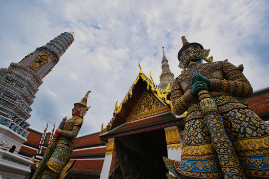 View of the ornate demon guardian statues at The Grand Palace with traditional Thai architecture and intricate golden details under a cloudy sky in Bangkok Thailand. View of the ornate demon guardian statues at The Grand Palace with traditional Thai architecture and intricate golden details under a cloudy sky in Bangkok Thailand.