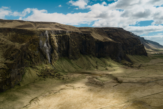 Aerial view of Seljalandsfoss waterfall cascading down a dramatic cliff face in a vast grassy landscape under a bright blue sky with scattered clouds in Rangarthing eystra Iceland. Aerial view of Seljalandsfoss waterfall cascading down a dramatic cliff face in a vast grassy landscape under a bright blue sky with scattered clouds in Rangarthing eystra Iceland.