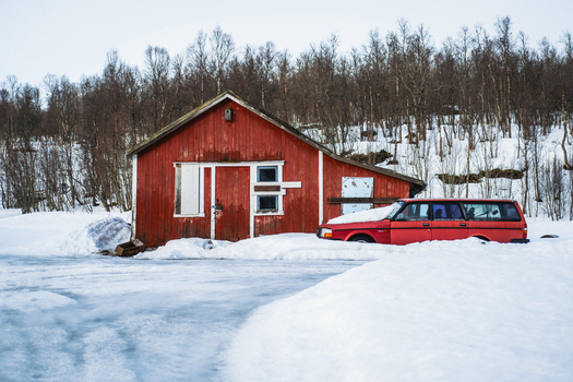 View of a red barn stands boldly against the stark white snow a matching red car parked nearby under the watchful gaze of bare trees Bardufoss Troms Norway. View of a red barn stands boldly against the stark white snow a matching red car parked nearby under the watchful gaze of bare trees Bardufoss Troms Norway.