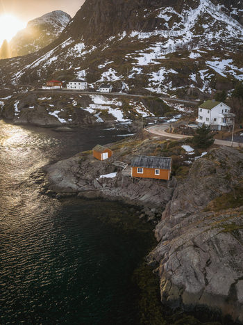 Aerial view of quaint orange cabins nestled on rocky outcrops where land meets the dark tranquil sea beneath snow dusted mountains Tind Nordland Norway.