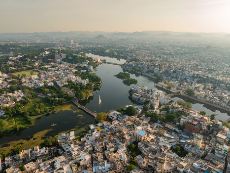Aerial view of the historic city of Udaipur with its dense urban architecture bridges and lush greenery surrounding the calm waters of Lake Pichola in Udaipur Rajasthan India.