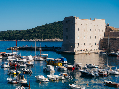 View of St. John s Fortress and the old harbor with small boats moored in the blue water of the Adriatic Sea and people on the pier in Dubrovnik Dubrovnik Neretva County Croatia. View of St. John s Fortress and the old harbor with small boats moored in the blue water of the Adriatic Sea and people on the pier in Dubrovnik Dubrovnik Neretva County Croatia.