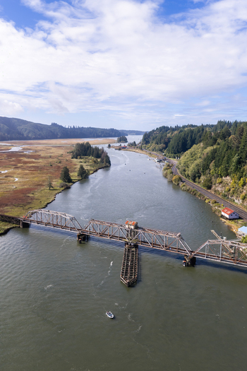 Aerial view of the historic Siuslaw River Train Bridge spanning the river surrounded by lush forested hills and wetlands with a small boat on the water Florence Oregon United States.