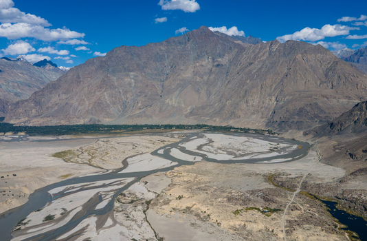 Aerial view of Shigar Valley featuring the braided Shigar River flowing through a vast sandy riverbed surrounded by rugged mountain peaks under a clear blue sky in Shigar Gilgit Pakistan.