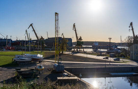 View of the industrial port area with large yellow cranes docked sailboats on trailers and a cargo ship in the background under a clear sky Klaipeda Klaipeda County Lithuania. View of the industrial port area with large yellow cranes docked sailboats on trailers and a cargo ship in the background under a clear sky Klaipeda Klaipeda County Lithuania.