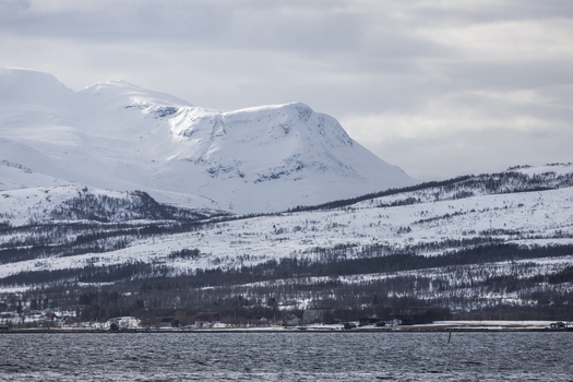View of snow covered mountain peaks overlooking a quiet coastal settlement and the dark waters of a fjord Tromso Troms Norway.