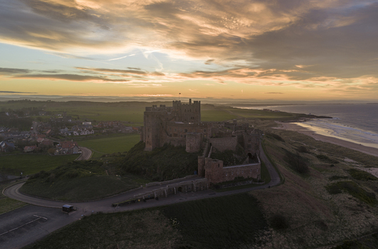 Aerial view of Bamburgh Castle perched on a rocky outcrop overlooking the North Sea coast and sandy beach under a dramatic sunset sky Bamburgh England United Kingdom. Aerial view of Bamburgh Castle perched on a rocky outcrop overlooking the North Sea coast and sandy beach under a dramatic sunset sky Bamburgh England United Kingdom.