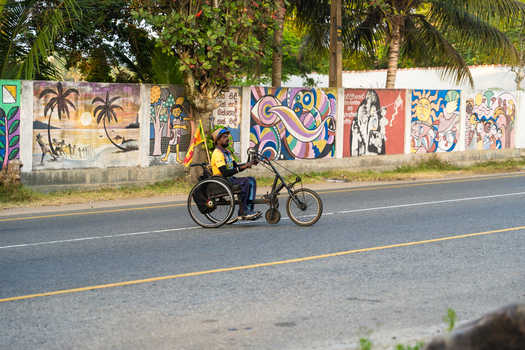 Sri Lanka 25 January 2026 View of a man riding a hand powered tricycle along a road past a long wall decorated with colorful murals and tropical trees.