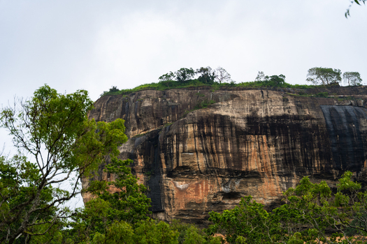 View of the massive granite cliffs of Sigiriya Rock Fortress surrounded by lush green trees under an overcast sky in Sigiriya Central Province Sri Lanka. View of the massive granite cliffs of Sigiriya Rock Fortress surrounded by lush green trees under an overcast sky in Sigiriya Central Province Sri Lanka.