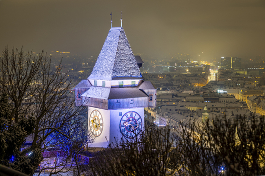 View of the iconic Grazer Uhrturm clock tower illuminated against the night sky overlooking a snow dusted cityscape twinkling below Graz Styria Austria. View of the iconic Grazer Uhrturm clock tower illuminated against the night sky overlooking a snow dusted cityscape twinkling below Graz Styria Austria.