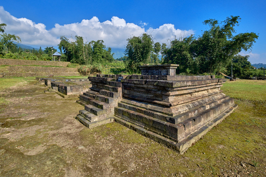 View of the ancient stone structures of Situs Liyangan archaeological site surrounded by lush greenery and trees under a blue sky in Temanggung Central Java Indonesia.