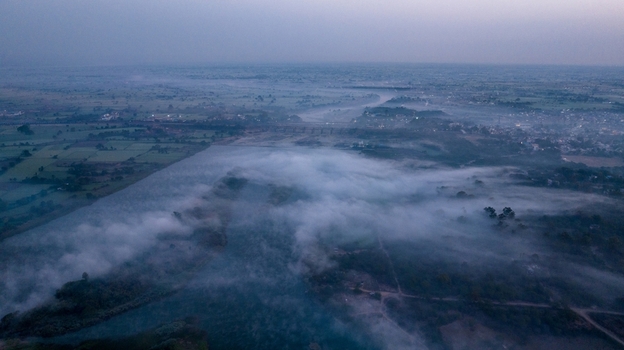Aerial view of the Chambal River flowing through the misty landscape with lush green fields and rural terrain under soft morning light in Nagda Madhya Pradesh India. Aerial view of the Chambal River flowing through the misty landscape with lush green fields and rural terrain under soft morning light in Nagda Madhya Pradesh India.