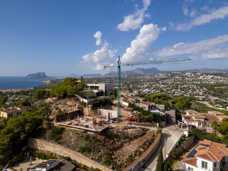 Aerial view of a construction site with a tall green crane on a hillside overlooking the coast surrounded by villas and greenery in Moraira Valencian Community Spain.