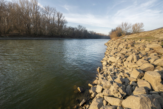 View of the rocky riverbank and calm water flowing past bare winter trees under a soft blue sky in Bratislava IV Bratislava Region Slovakia. View of the rocky riverbank and calm water flowing past bare winter trees under a soft blue sky in Bratislava IV Bratislava Region Slovakia.