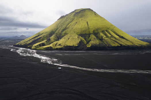 Aerial view of the verdant mossy Maelifell volcano rising in stark contrast from the jet black volcanic sands a silver river carving its path below Maelifell Iceland. Aerial view of the verdant mossy Maelifell volcano rising in stark contrast from the jet black volcanic sands a silver river carving its path below Maelifell Iceland.