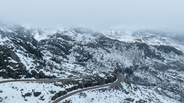 Aerial view of the winding mountain road through the snow covered peaks of the Serra da Estrela under a misty sky near the glacial valley deck Loriga Guarda District Portugal. Aerial view of the winding mountain road through the snow covered peaks of the Serra da Estrela under a misty sky near the glacial valley deck Loriga Guarda District Portugal.