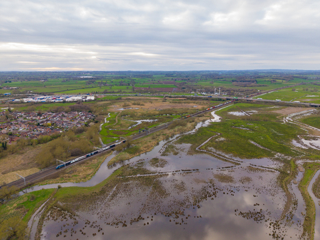 Aerial view of Doxey Marshes with a passenger train traveling along the tracks surrounded by wetlands green fields and suburban housing under a cloudy sky in Stafford England United Kingdom. Aerial view of Doxey Marshes with a passenger train traveling along the tracks surrounded by wetlands green fields and suburban housing under a cloudy sky in Stafford England United Kingdom.