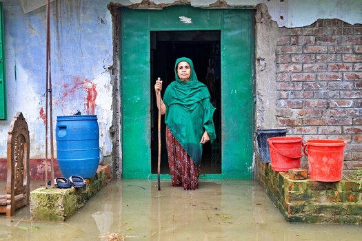 Banshkhali Upazila Chittagong Division Bangladesh 10 July 2026 View of an elderly woman standing in floodwater at the entrance of her home surrounded by plastic buckets. Banshkhali Upazila Chittagong Division Bangladesh 10 July 2026 View of an elderly woman standing in floodwater at the entrance of her home surrounded by plastic buckets.
