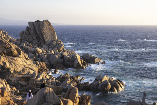 View of the massive granite boulders and rugged coastline with people overlooking the blue sea during golden hour at Capo Testa Sardegna Italy. View of the massive granite boulders and rugged coastline with people overlooking the blue sea during golden hour at Capo Testa Sardegna Italy.