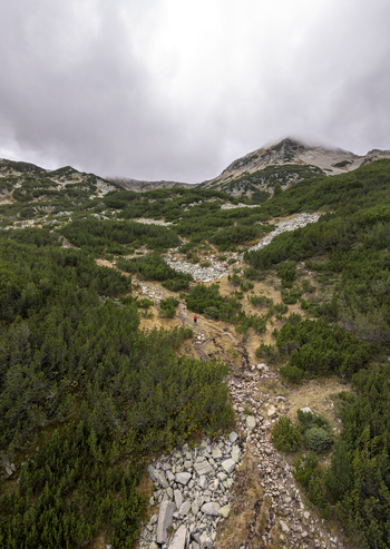 Aerial view of Pirin National Park with a hiker in a red jacket on a rocky path near Muratovo Lake surrounded by pine shrubs and mountain peaks in Blagoevgrad Province Bulgaria. Aerial view of Pirin National Park with a hiker in a red jacket on a rocky path near Muratovo Lake surrounded by pine shrubs and mountain peaks in Blagoevgrad Province Bulgaria.