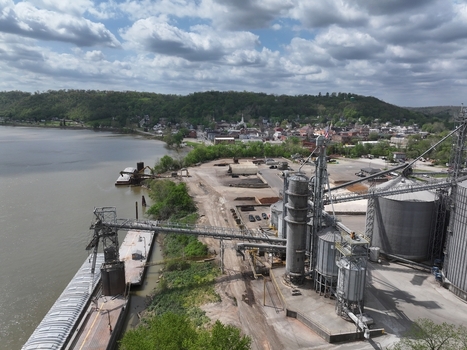 Aerial view of an industrial grain elevator facility and docked barges along the riverbank with the town skyline and green hills under a cloudy sky Aurora Indiana United States. Aerial view of an industrial grain elevator facility and docked barges along the riverbank with the town skyline and green hills under a cloudy sky Aurora Indiana United States.