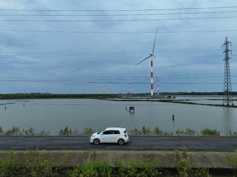 Aerial view of a white car driving along a road next to the Khurushkul wind farm with turbines in flooded fields under an overcast sky in Cox s Bazar Chittagong Division Bangladesh. Aerial view of a white car driving along a road next to the Khurushkul wind farm with turbines in flooded fields under an overcast sky in Cox s Bazar Chittagong Division Bangladesh.