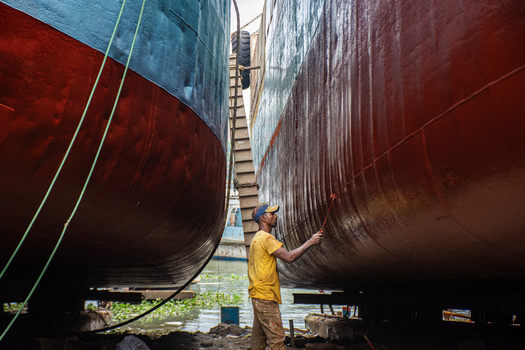 28 August 2022 View of a worker in a yellow shirt performing maintenance on a large ship hull in a shipyard.
