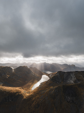 Aerial view of the rugged mountains and a reflective lake on Senja island under dramatic clouds with sun rays piercing through in Senja Troms Norway.