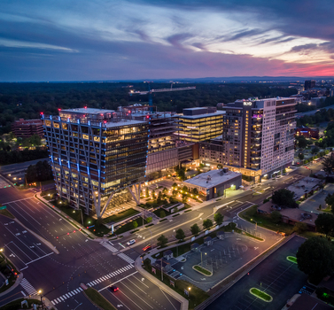 Reston Virginia United States 27 May 2020 Aerial view of modern office buildings and mixed use developments at twilight featuring illuminated glass facades a construction crane and surrounding roads. Reston Virginia United States 27 May 2020 Aerial view of modern office buildings and mixed use developments at twilight featuring illuminated glass facades a construction crane and surrounding roads.