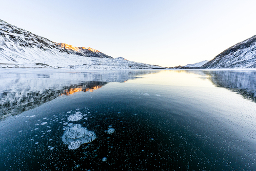 View of a frozen lake with methane bubbles trapped in the ice surrounded by snow covered mountains under a clear sky at sunset Madesimo Lombardy Italy. View of a frozen lake with methane bubbles trapped in the ice surrounded by snow covered mountains under a clear sky at sunset Madesimo Lombardy Italy.