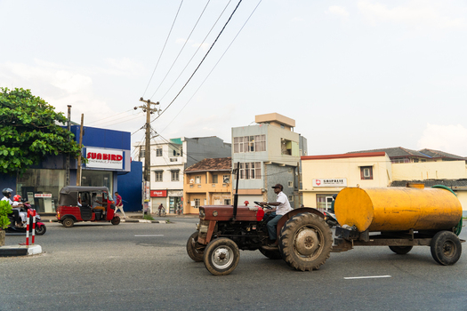 Sri Lanka 15 January 2026 View of a red tractor pulling a yellow water tank trailer along a busy urban street with a driver a tuk tuk and commercial buildings.