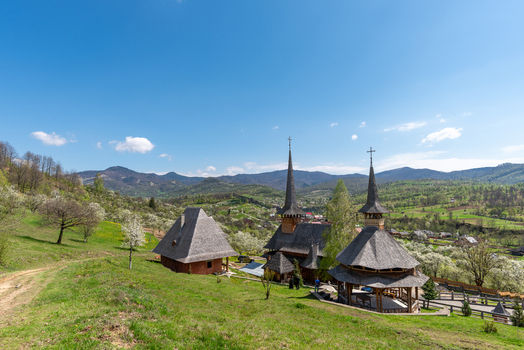View of a traditional wooden church with tall spires surrounded by blooming trees and green hills in Maramures Romania. View of a traditional wooden church with tall spires surrounded by blooming trees and green hills in Maramures Romania.