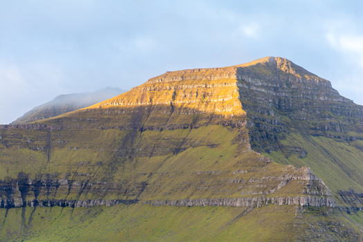 View of a rugged mountain peak with layered rock formations and vibrant green slopes illuminated by golden hour sunlight in the Faroe Islands. View of a rugged mountain peak with layered rock formations and vibrant green slopes illuminated by golden hour sunlight in the Faroe Islands.