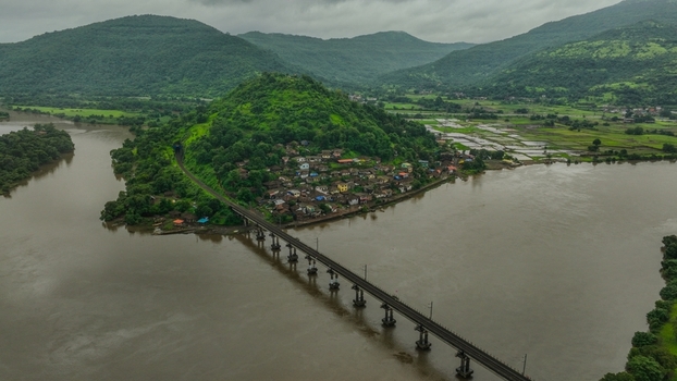 Aerial view of a railway bridge crossing a wide river with a small village nestled on a green hill under a cloudy sky in Mahad Maharashtra India.