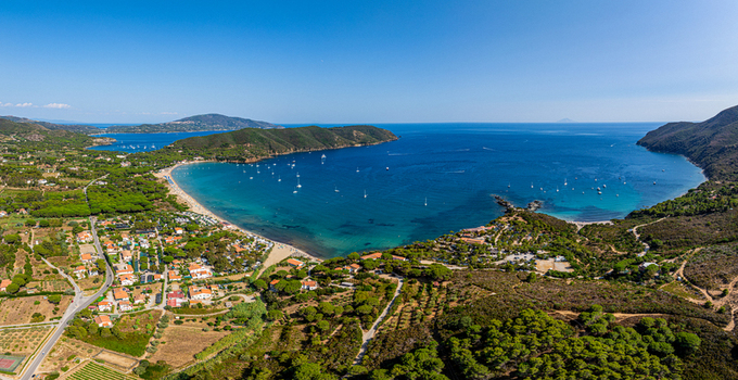 Aerial view of Lacona Beach with turquoise waters sandy coastline anchored boats and surrounding green hills under a clear blue sky in Lacona Tuscany Italy.