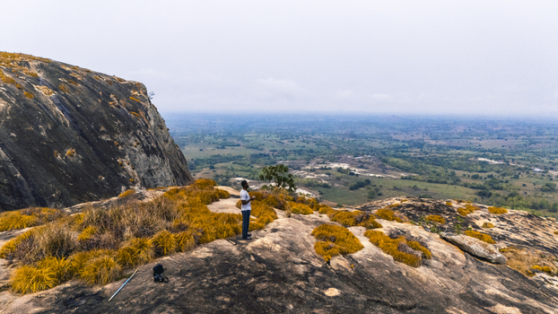 Aerial view of a figure standing atop a rocky outcrop overlooking the vast IYAKE LAKE where brown grasses meet rugged stone Oyo Oyo Nigeria. Aerial view of a figure standing atop a rocky outcrop overlooking the vast IYAKE LAKE where brown grasses meet rugged stone Oyo Oyo Nigeria.
