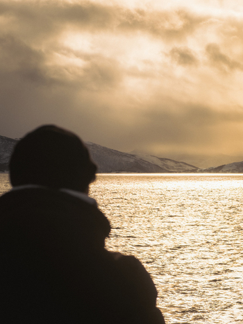 View of a silhouetted figure gazing out at the shimmering sea reflecting the golden sky framed by distant snow capped mountains Brensholmen Norway. View of a silhouetted figure gazing out at the shimmering sea reflecting the golden sky framed by distant snow capped mountains Brensholmen Norway.