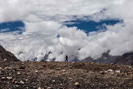 View of a hiker with a green backpack trekking across the rocky moraine of the Baltoro Glacier under a dramatic cloudy sky in Baltoro Gilgit Baltistan Pakistan.