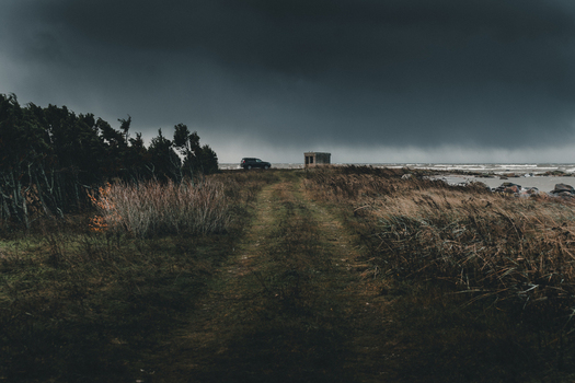 View of Vormsi island landscape with a dirt path leading to a small structure and a parked car under dramatic dark storm clouds in Laane County Estonia.